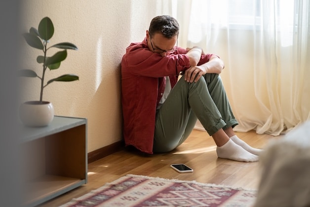 Depressed man sits on floor looking at mobile phone waiting for call from treatment center.